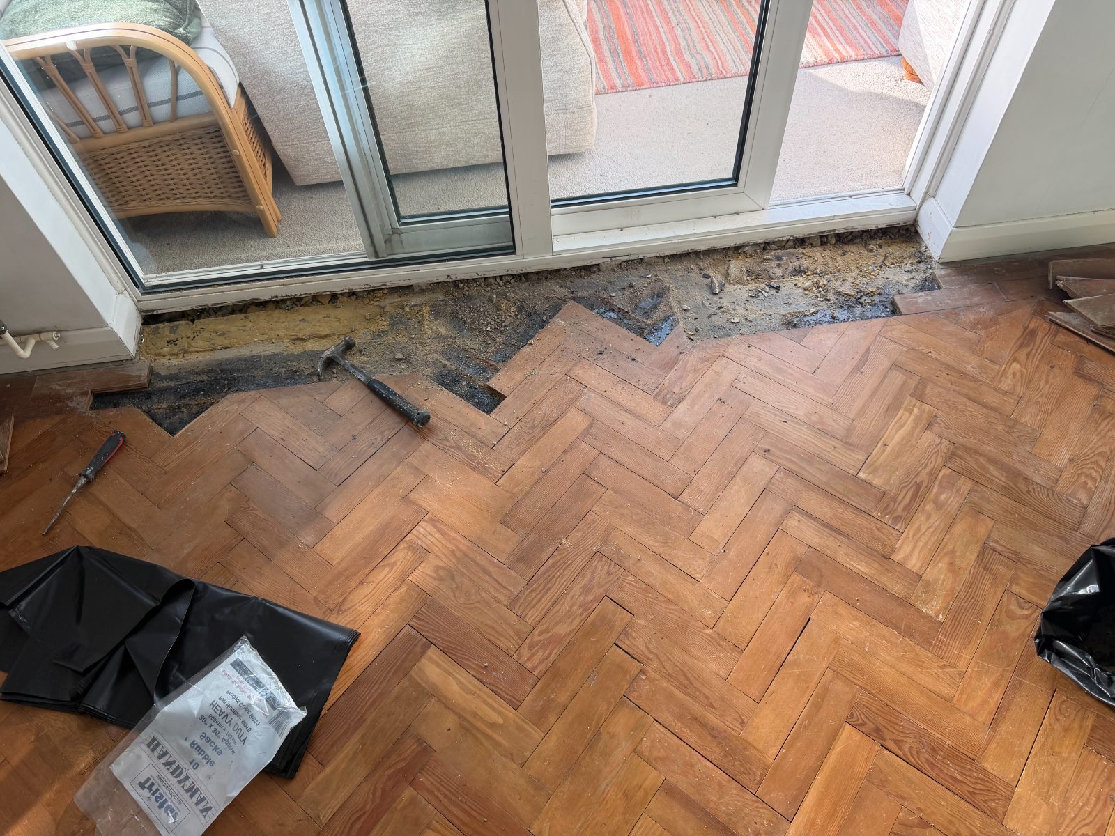 Pitch-pine herringbone parquet mid-repair at patio doors, blocks lifted and damaged subfloor exposed, hammer and chisel on the floor