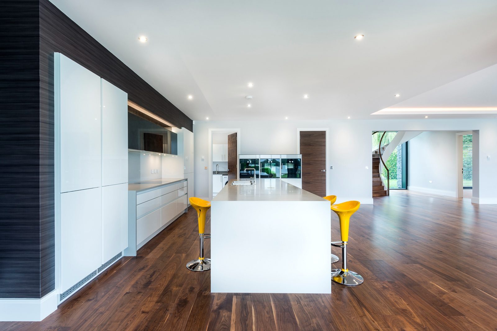 Rich dark walnut engineered plank flooring running through a contemporary open-plan kitchen with a white island, leading toward a curved staircase in a Birdham home