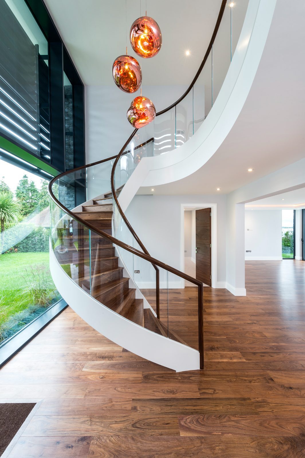 A curved feature staircase clad in matching walnut with a glass balustrade and copper pendant lights, rising from a walnut plank floor in a Birdham new-build