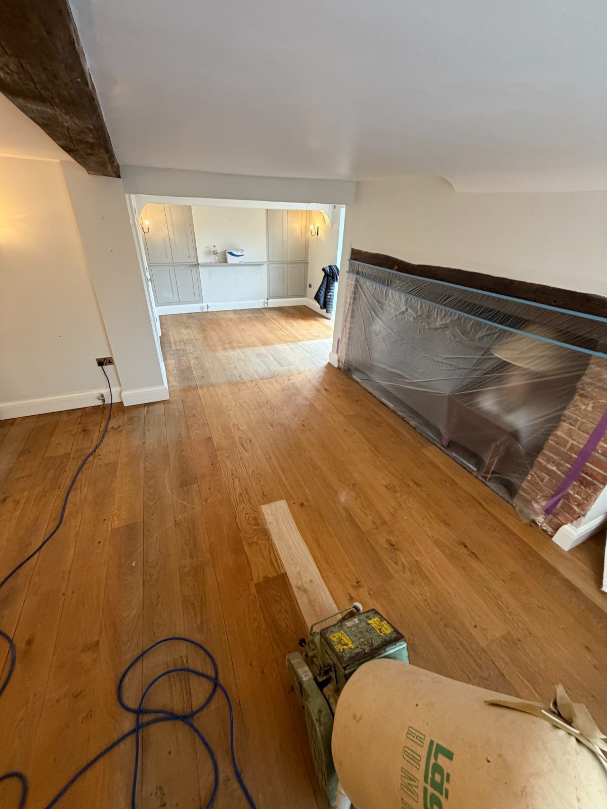 Worn solid oak wide-plank flooring being sanded back to bare timber with a belt sander in a beamed Slindon living room, restoration in progress