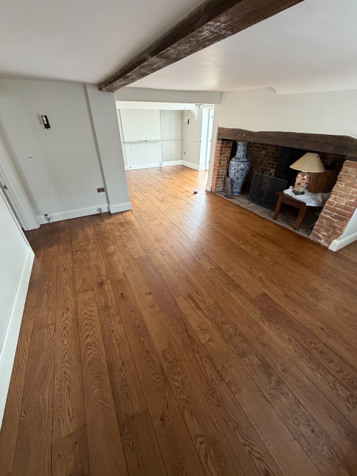Restored solid oak wide-plank flooring finished in a rich dark walnut stain and matt hard-wax oil, sweeping across a beamed period living room with an inglenook brick fireplace in a Slindon cottage