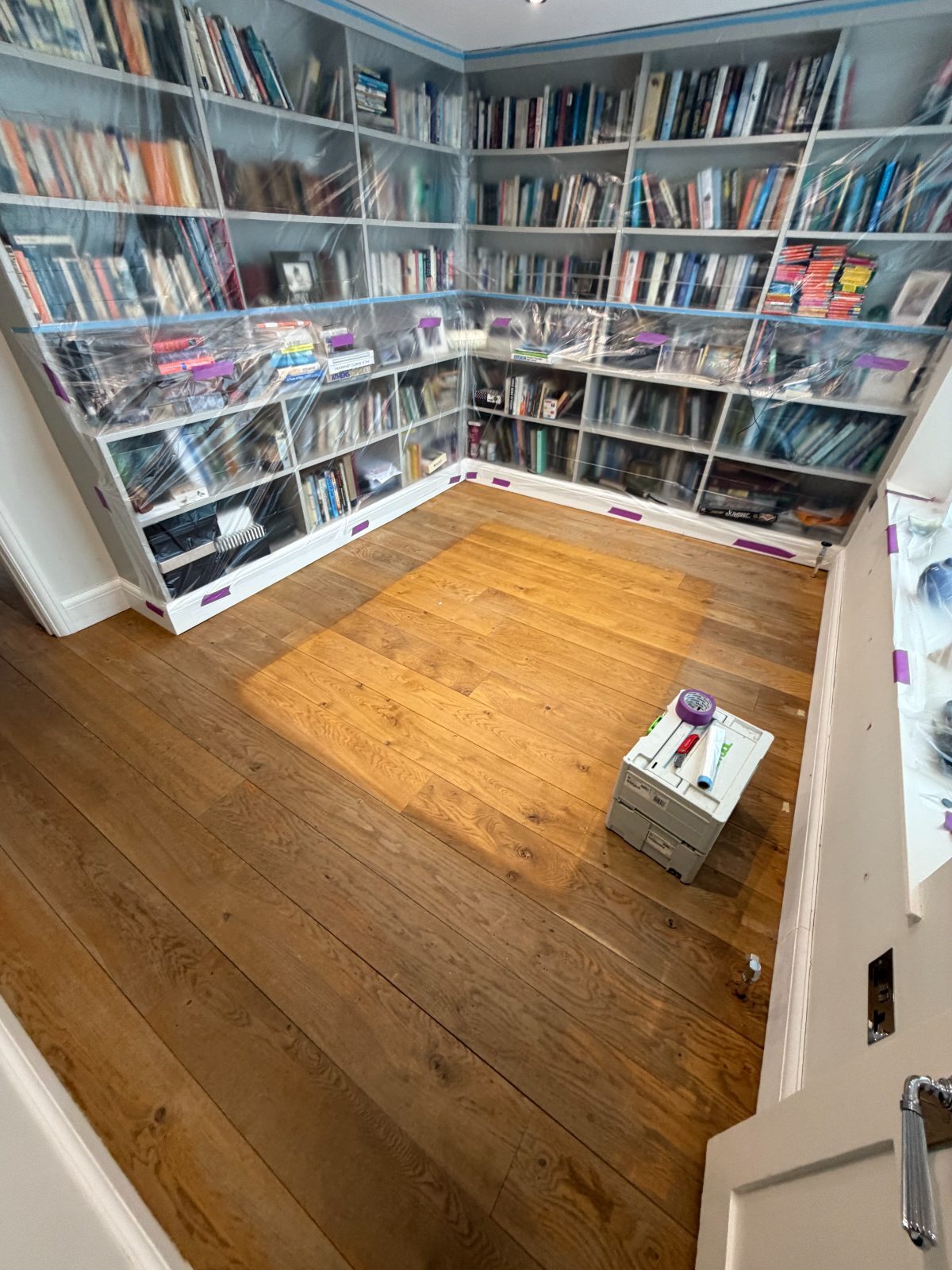 Worn solid oak wide-plank flooring being sanded back to bare timber in a Slindon period library, fitted bookcases sheeted in plastic for dust protection, restoration in progress