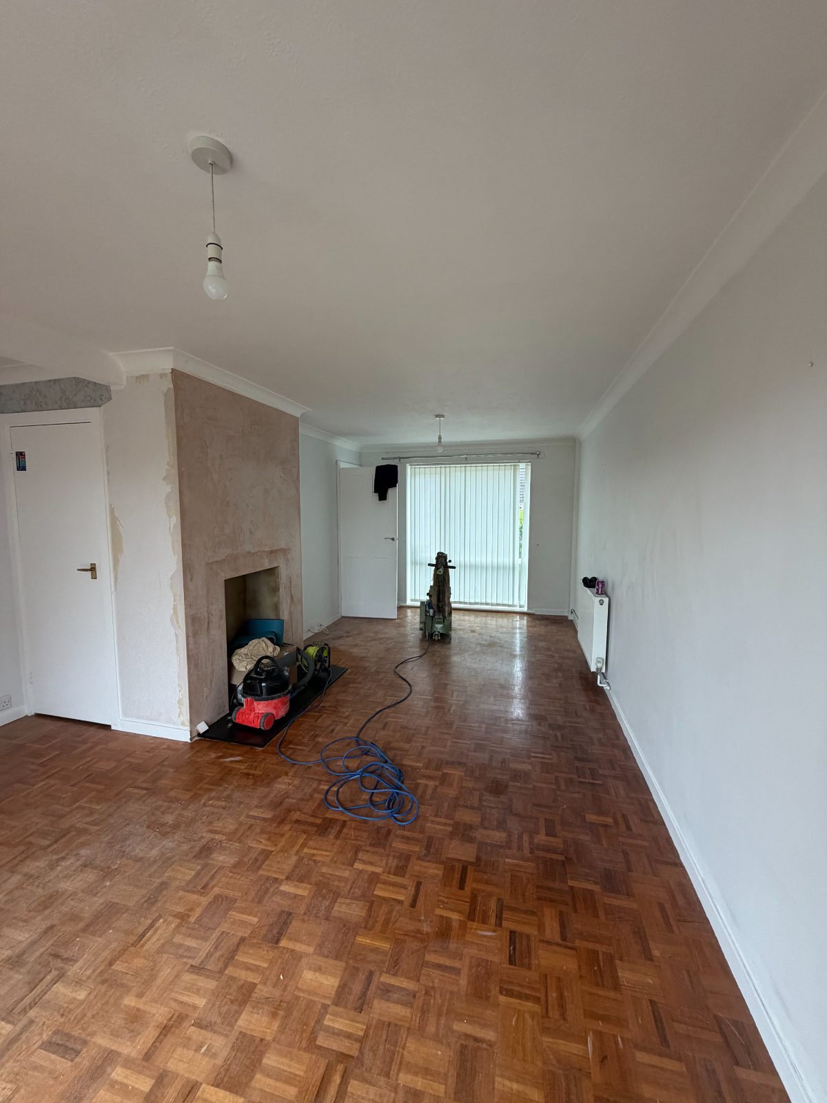 An original 1960s mosaic-block parquet floor part-way through restoration in a Westergate living room, the floor sander still on the boards and the chimney breast stripped back to plaster