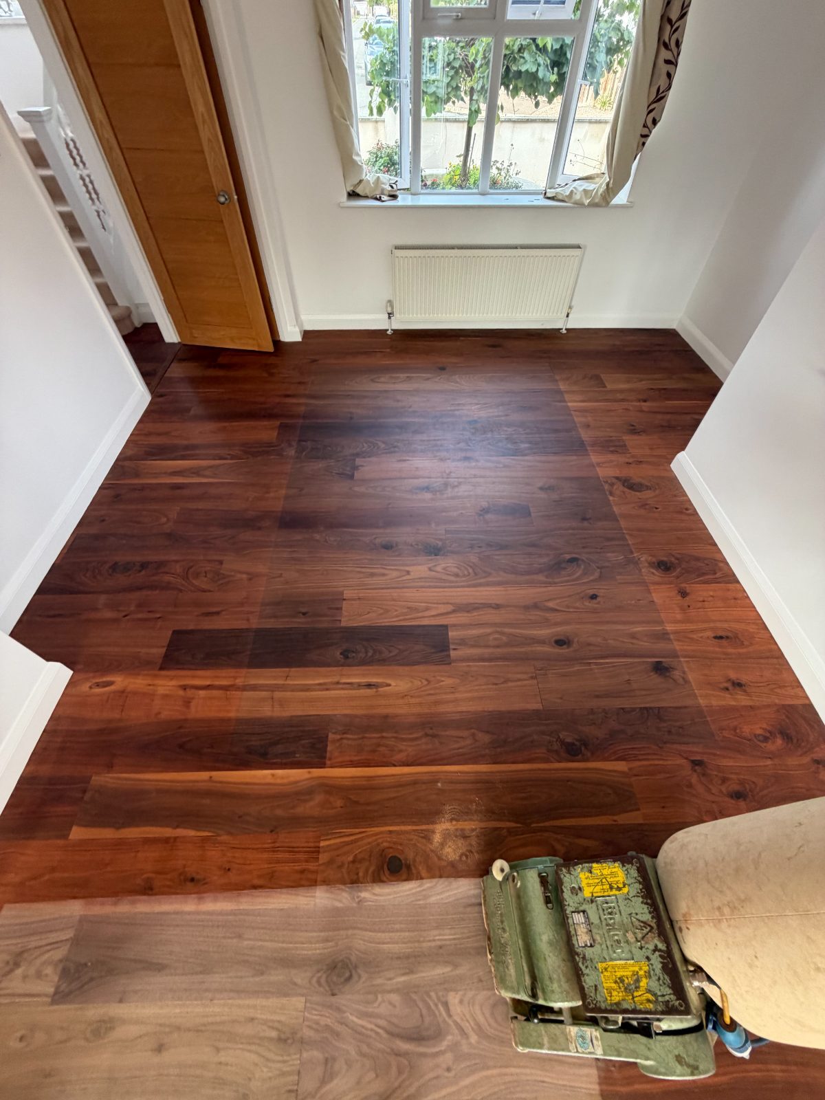 A wide-plank walnut floor mid-sand in a Felpham hallway, the drum sander halfway across with bare sanded boards against the old darker finish
