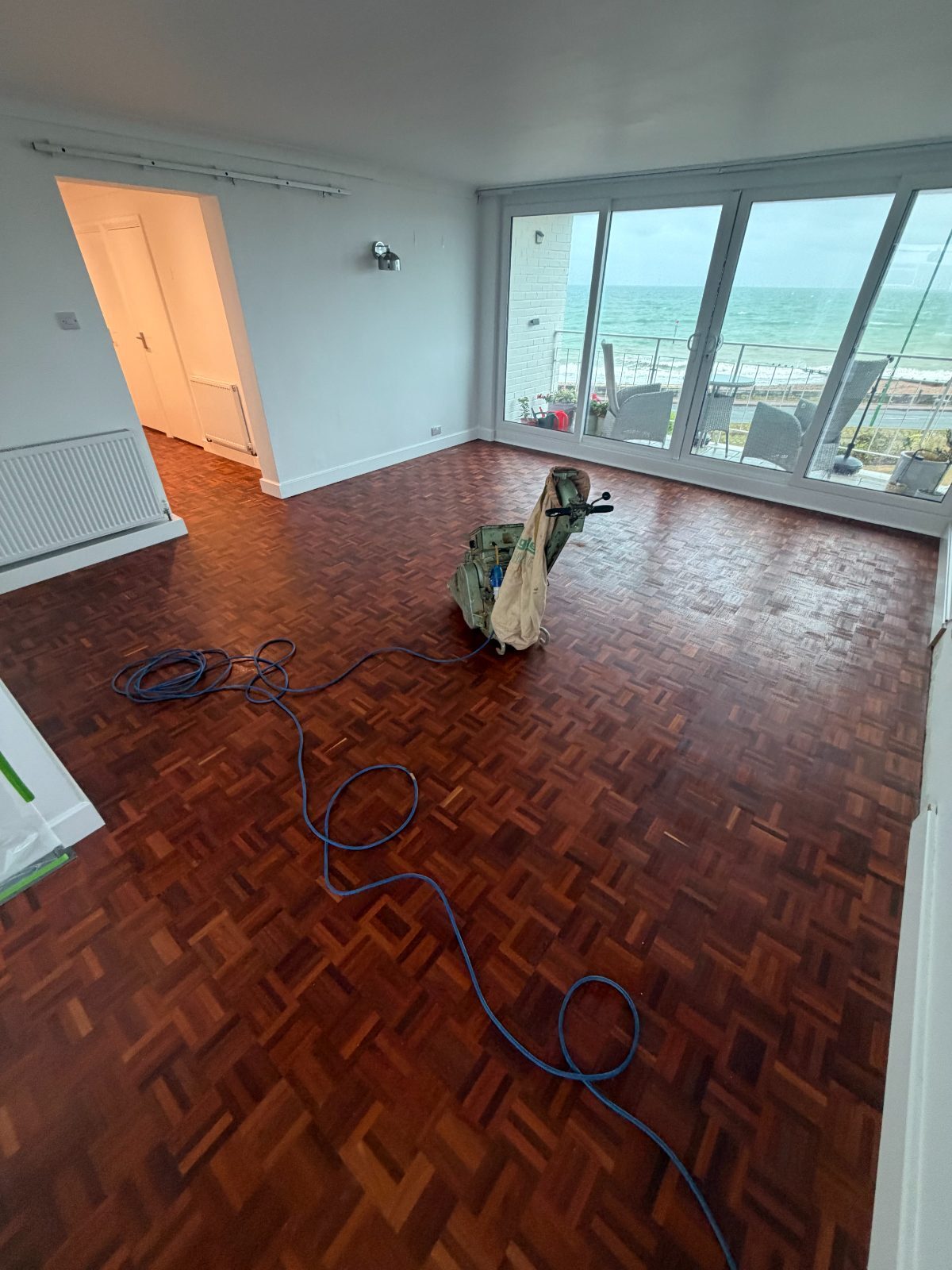 A mosaic parquet floor halfway through restoration in a Rustington seafront flat, the belt sander on the old dark finish and the sea through the balcony glazing