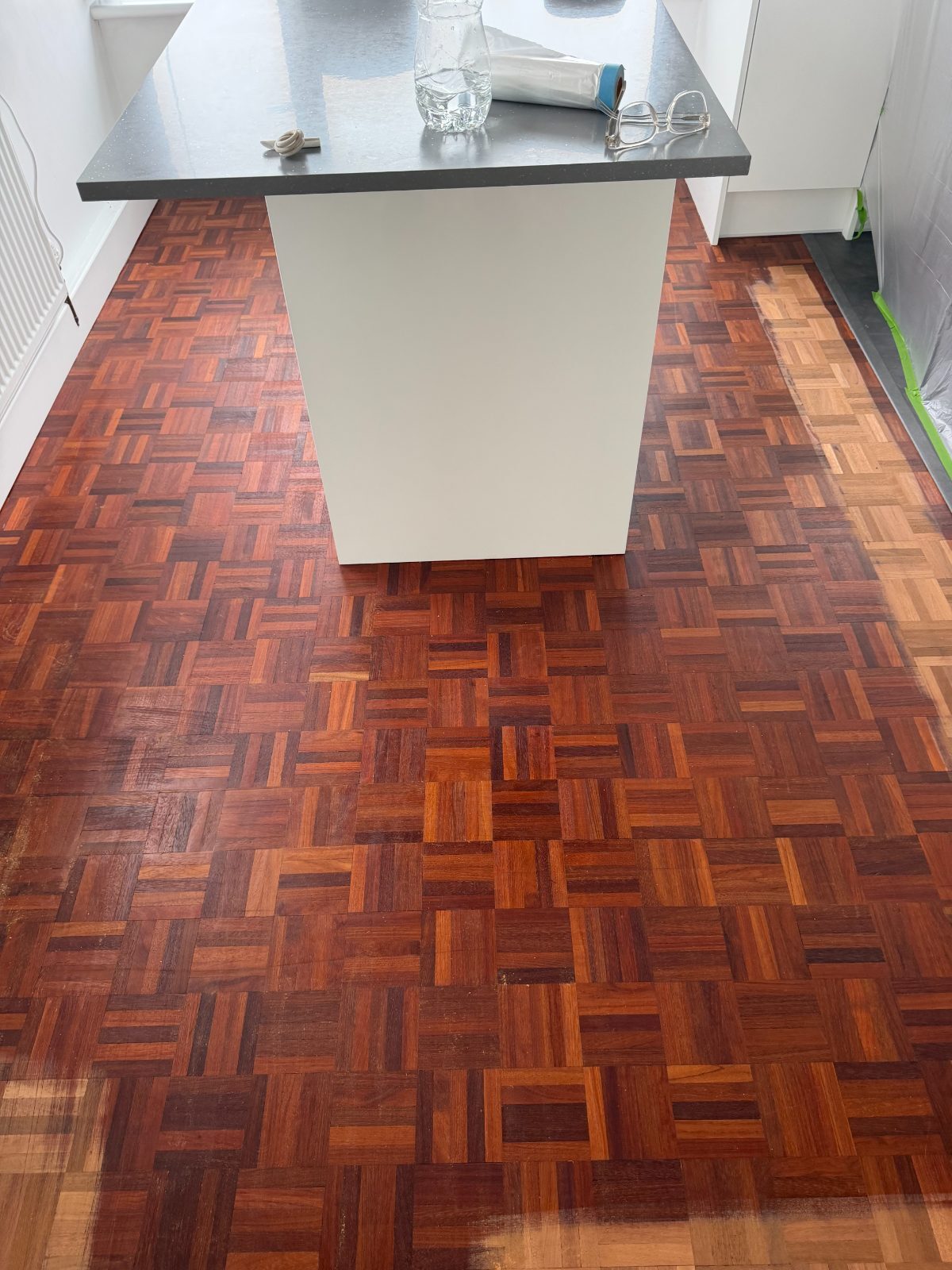 Mosaic parquet around a white kitchen island in a Rustington flat, part-sanded with the old dark finish still on the centre and pale bare timber at the edges