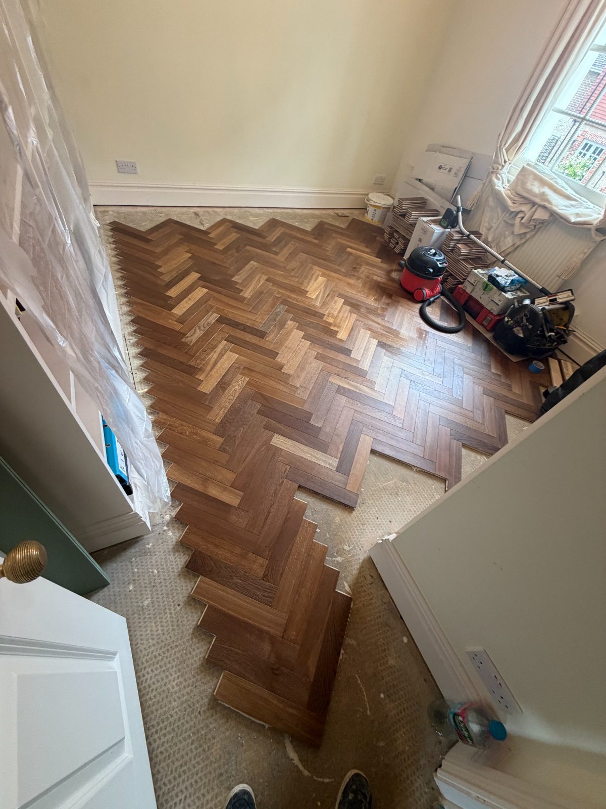 Overhead view of oak herringbone parquet part-laid across a sash-windowed room at Burton House, tools and dust sheets around the edges